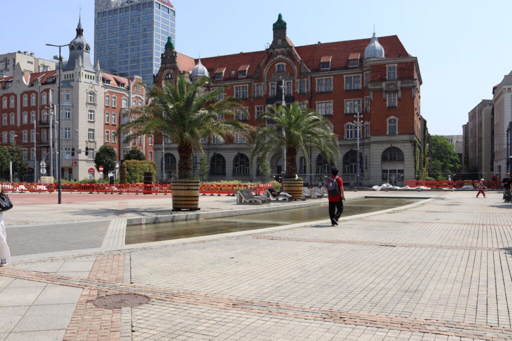 Widok na historyczny budynek i nowoczesny wieżowiec przy rynku Katowic. Na pierwszym planie sztucznie poprowadzona rzeka rawa i dwie palmy oraz ławki. Spaceruje kilka osób.