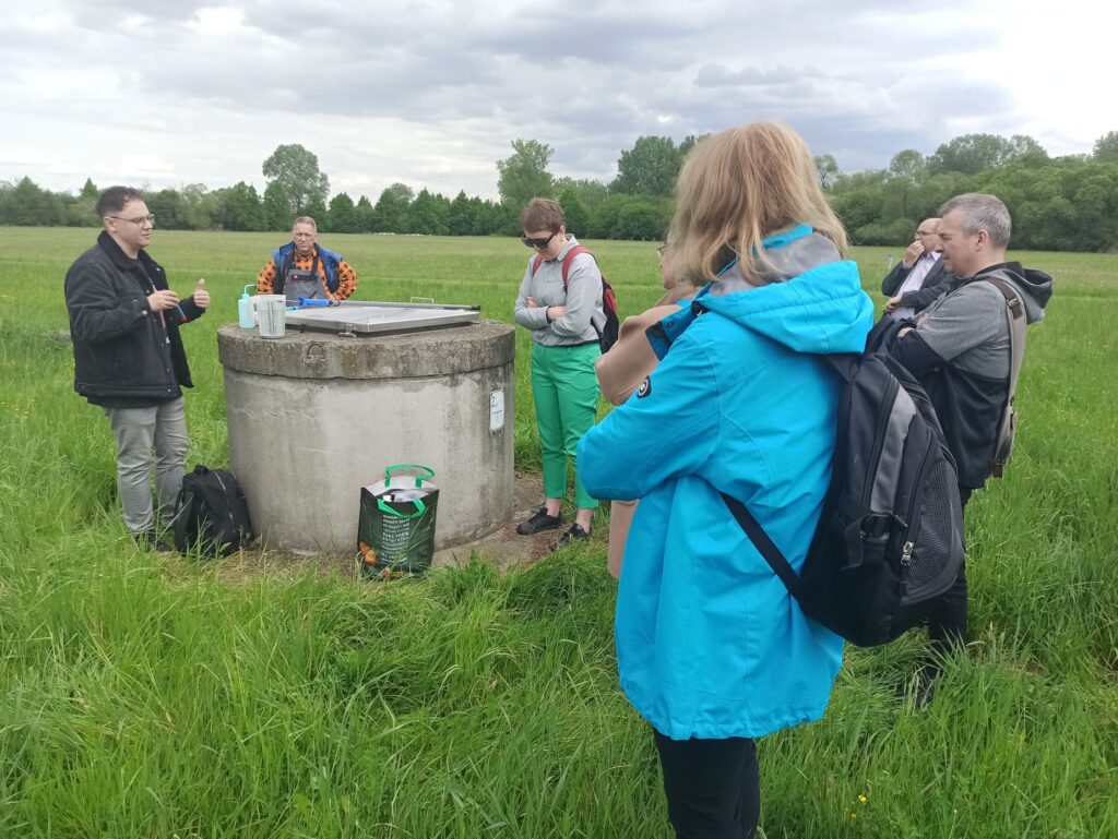 Grupa uczestników zgromadzona wokół ujęcia filtracyjnego wody podczas prezentacji sprzętu do pomiarów hydrogeologicznych.