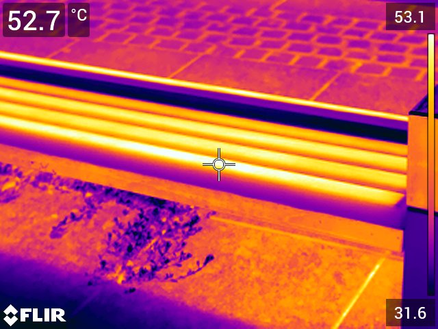 Termowizyjne zbliżenie na drewnianą ławkę miejską. Siedzisko ma bardzo wysoką temperaturę powierzchni, przekraczającą 50°C, podczas gdy otaczające je elementy są wyraźnie chłodniejsze.