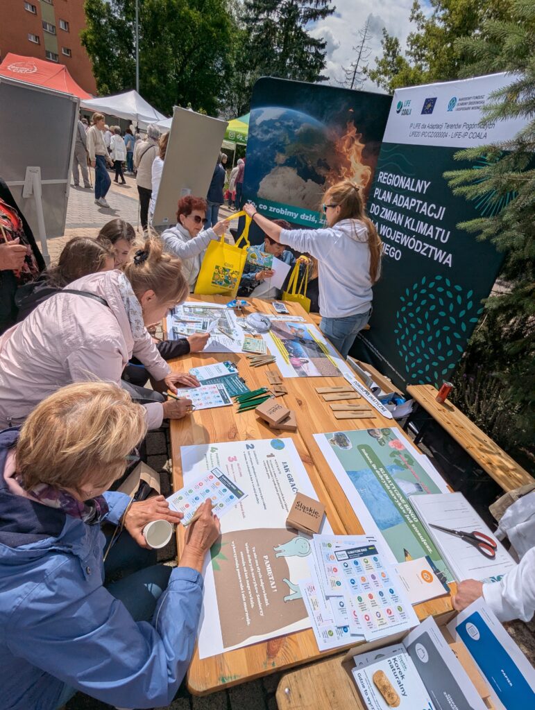 Seniorzy biorą udział w warsztatach edukacyjnych na temat zmian klimatu przy plenerowym stoisku informacyjnym.