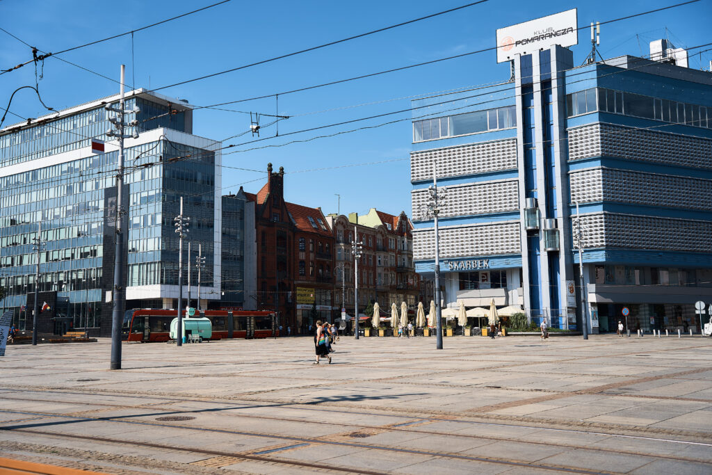 Widok na Rynek w Katowicach w słoneczny dzień. Po lewej stronie widoczny jest tramwaj jadący po torowisku, a przez plac spaceruje kilka osób. Po prawej stronie znajdują się charakterystyczny budynek Skarbka oraz sąsiadujące kamienice przy ulicy 3 Maja. W centrum kadru rozciąga się betonowa przestrzeń rynku, nad którą rozpościera się błękitne, bezchmurne niebo.