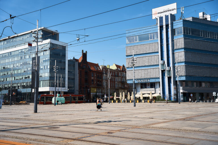 Widok na Rynek w Katowicach w słoneczny dzień. Po lewej stronie widoczny jest tramwaj jadący po torowisku, a przez plac spaceruje kilka osób. Po prawej stronie znajdują się charakterystyczny budynek Skarbka oraz sąsiadujące kamienice przy ulicy 3 Maja. W centrum kadru rozciąga się betonowa przestrzeń rynku, nad którą rozpościera się błękitne, bezchmurne niebo.