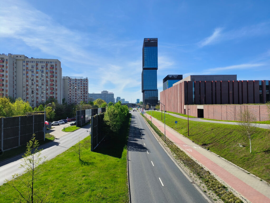 Nowoczesna zabudowa Katowic z wieżowcem biurowym, szeroką arterią komunikacyjną, ścieżką rowerową oraz terenami zielonymi w słoneczny dzień.