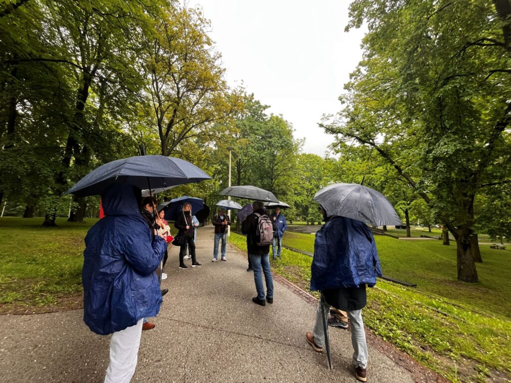 Grupa osób stoi na parkowej alejce podczas deszczu. Większość uczestników trzyma parasole, a dwie osoby mają niebieskie peleryny przeciwdeszczowe. Wokół rosną gęste, zielone drzewa, a mokra nawierzchnia i pochmurne niebo podkreślają deszczową pogodę.