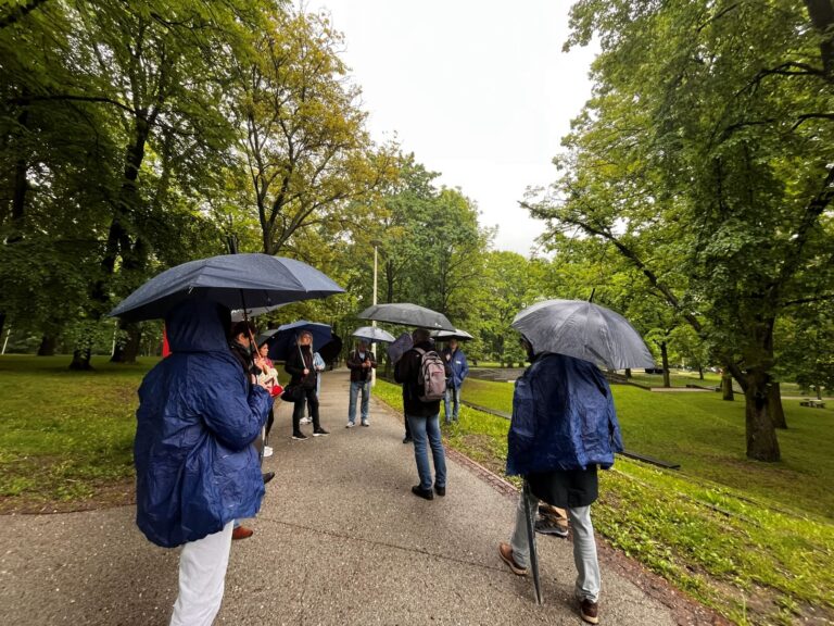 Grupa osób stoi na parkowej alejce podczas deszczu. Większość uczestników trzyma parasole, a dwie osoby mają niebieskie peleryny przeciwdeszczowe. Wokół rosną gęste, zielone drzewa, a mokra nawierzchnia i pochmurne niebo podkreślają deszczową pogodę.