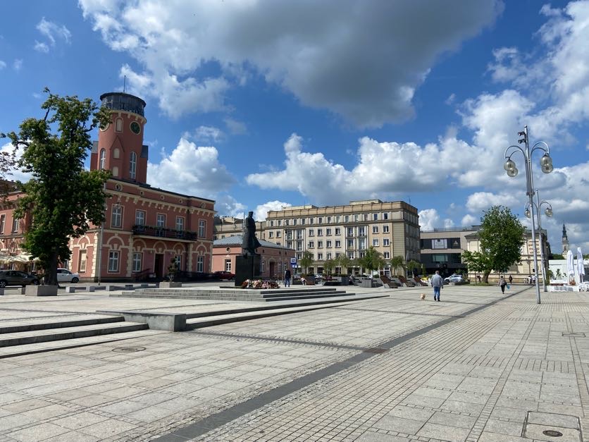 Widok na plac Biegańskiego w Częstochowie z rozległą utwardzoną nawierzchnią, zabytkowym budynkiem z wieżą oraz niewielką liczbą drzew i pieszych.